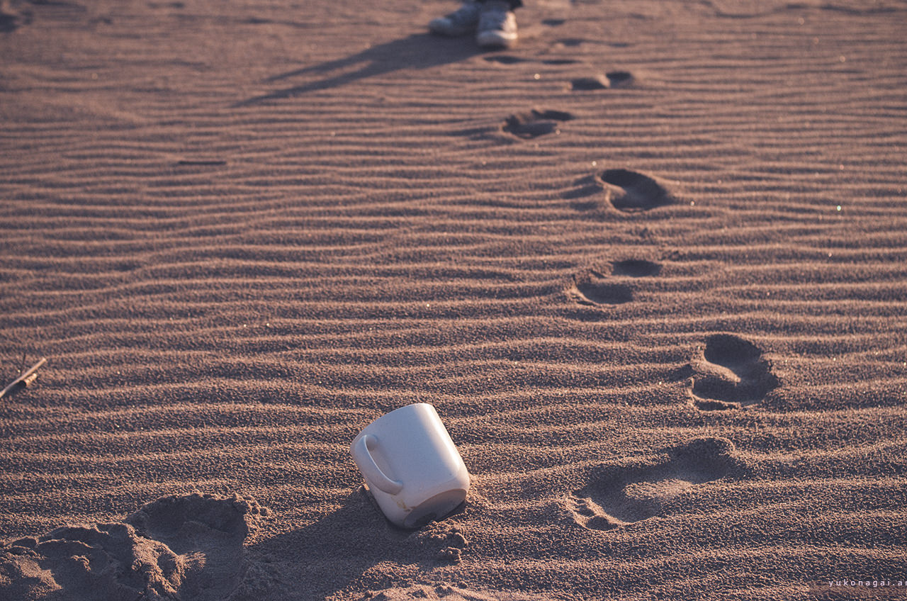 A mug cup and a pair of tennis shoes on beach sand ripples with foot prints.