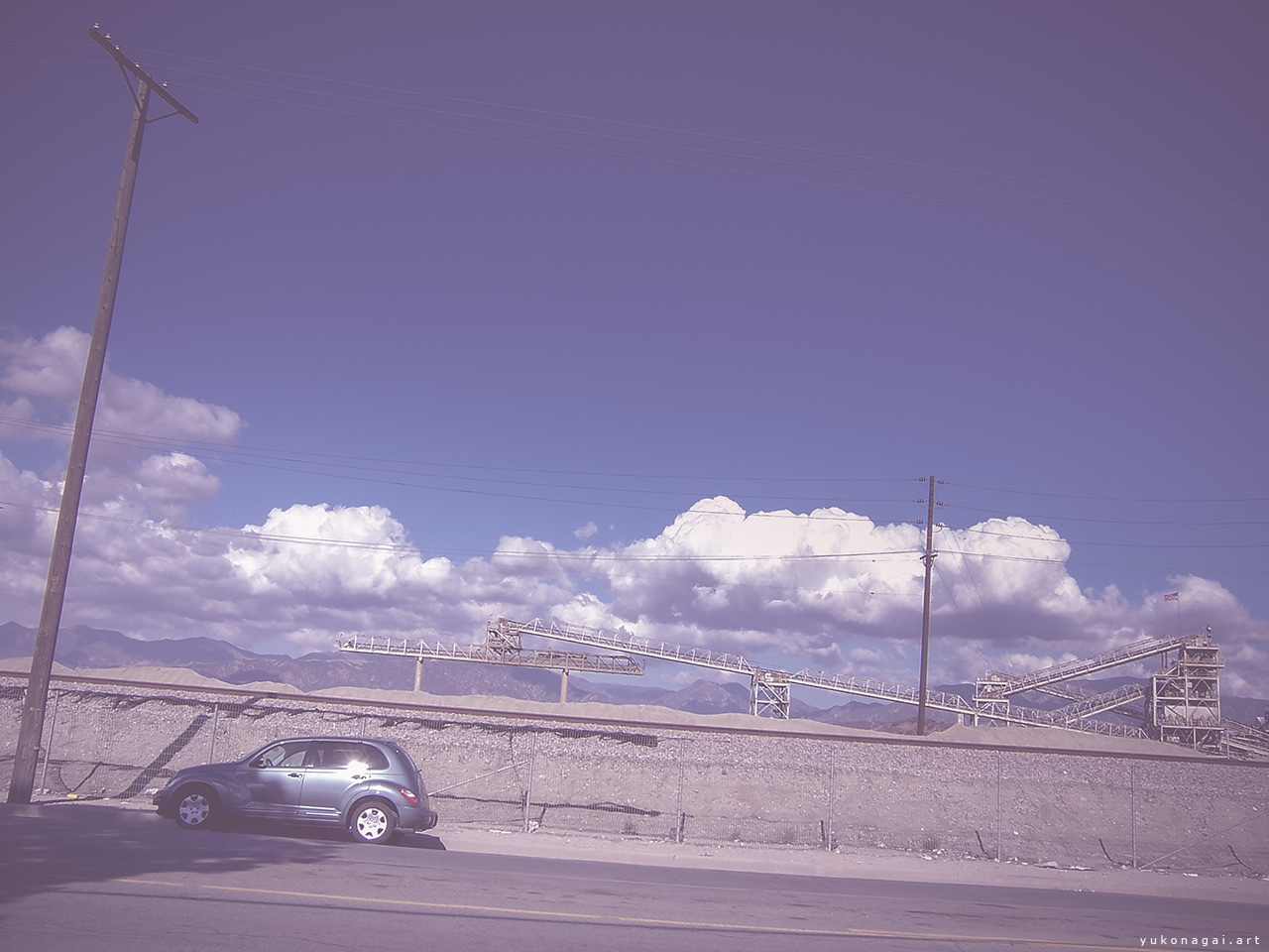 A silver car parked with mountains, clouds and concrete factory in background.